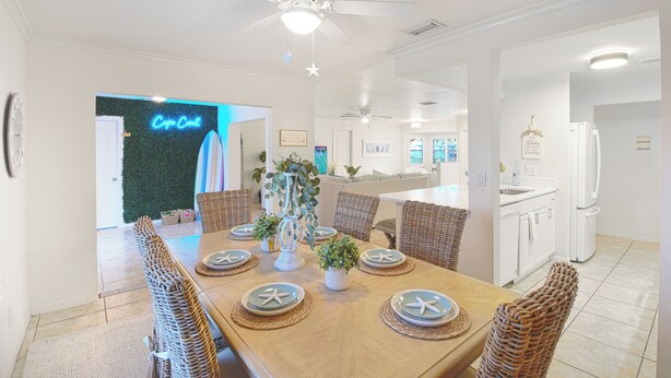 A dining area with a wooden table set for six, surrounded by wicker chairs. There is a ceiling fan above and a kitchen visible in the background.