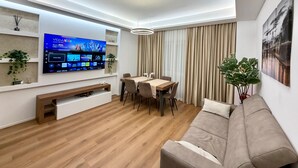 A modern living room with a flat-screen TV mounted on the wall, a dining area with a wooden table and chairs, a sofa, and a ceiling light fixture. - House Gordiani Comfort Stay (Roma)
