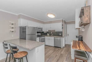 A kitchen with white cabinets, a granite countertop, stainless steel appliances, and a wooden dining table. - Luxury & Updated 2BR w/ Ocean views! (Horry County)