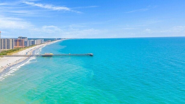 A coastal view with a pier extending into turquoise waters, a beach, and high-rise buildings in the background.