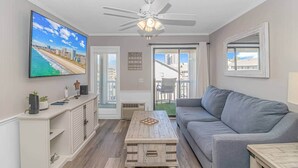 A living room with a flat-screen TV, a ceiling fan, a gray sofa, a wooden coffee table, and a view of the city through the balcony doors.