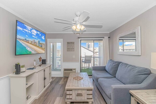 A living room with a flat-screen TV, a ceiling fan, a gray sofa, a wooden coffee table, and a view of the city through the balcony doors.