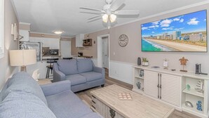 A living room with a gray sofa, a coffee table, a TV displaying a beach scene, a ceiling fan, and a kitchen area in the background.