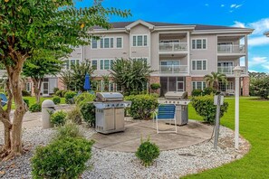 A multi-story building with a patio area featuring a grill, a barbecue, and a trash can.