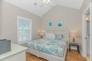 A bedroom with a bed, two bedside tables, a window with blinds, a ceiling fan, and a door leading to another room.