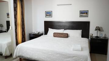 Een netjes opgemaakt bed met een witte spreideken en een bruin kussen, twee nachtkastjes met lampen, en twee ingekaderde foto’s aan de muur.