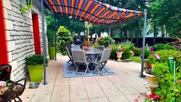 Une terrasse avec une table et des chaises, entourée de plantes en pot et d’un auvent rayé aux couleurs vives.