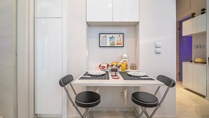 A compact dining area with a white table, two black chairs, a framed picture on the wall, and a small kitchenette with a purple accent wall. - Cool Style Private Apartments (Dafni-Ymittos)