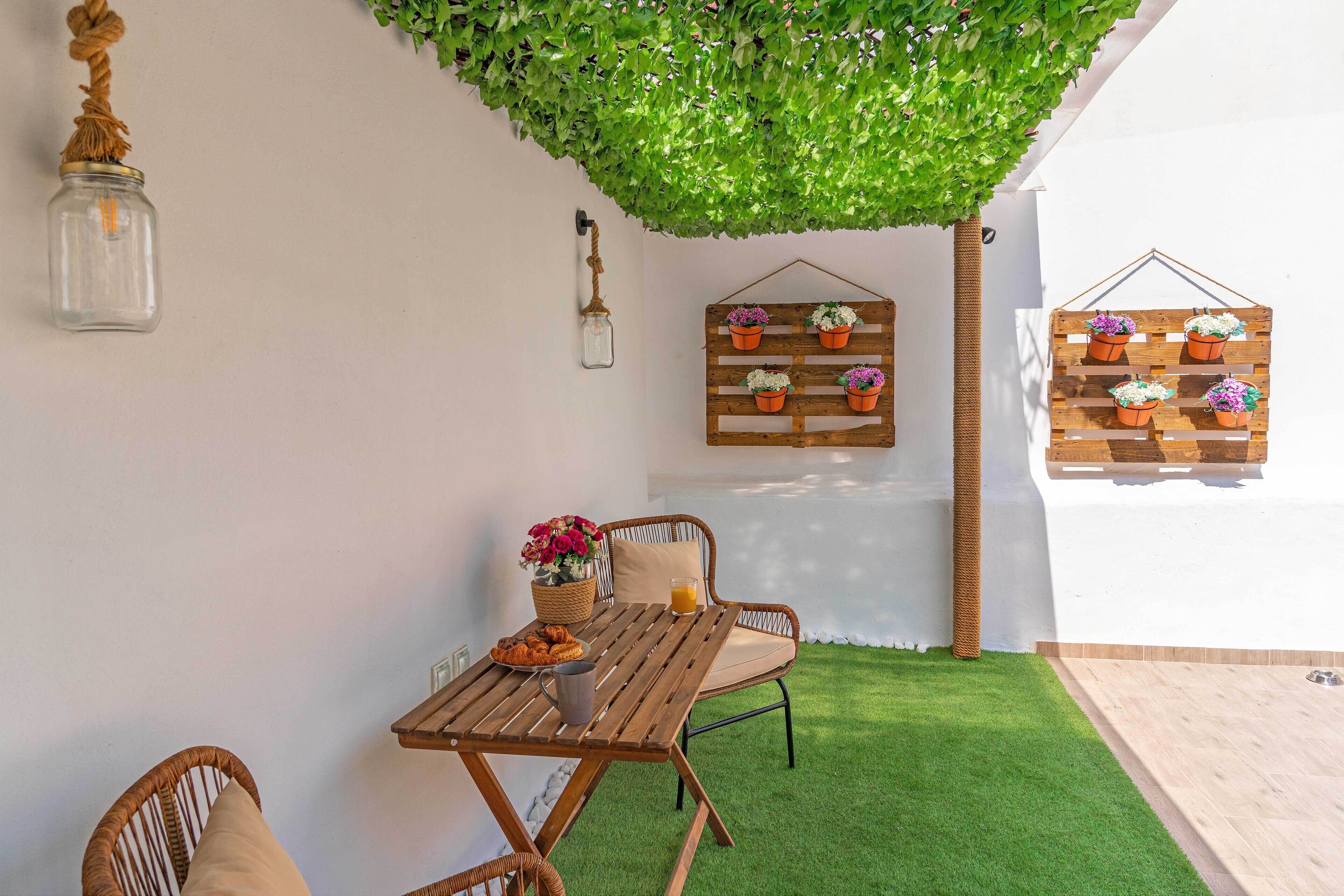 Una zona de estar al aire libre acogedora con mesa de madera y sillas de mimbre, rodeada de césped artificial exuberante y plantas en macetas decorativas.