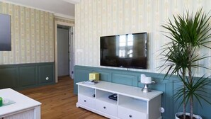 A living room with a flat-screen TV mounted on the wall, a white entertainment unit below, a potted plant, and a door leading to another room.