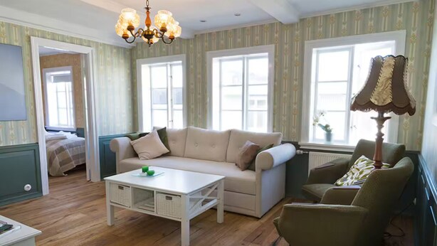 A cozy living room with a sofa, a coffee table, and a lamp. There are windows and a door visible.