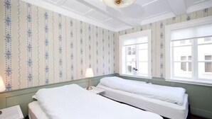 A hotel room with two single beds, a nightstand with a lamp, and a window with a view of buildings.