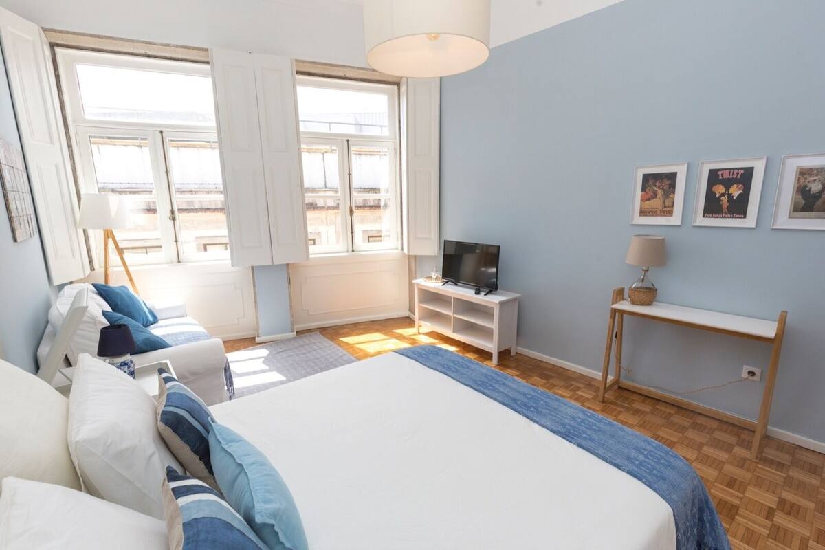 Une chambre avec un grand lit, un téléviseur posé sur un meuble blanc et une fenêtre avec des rideaux.