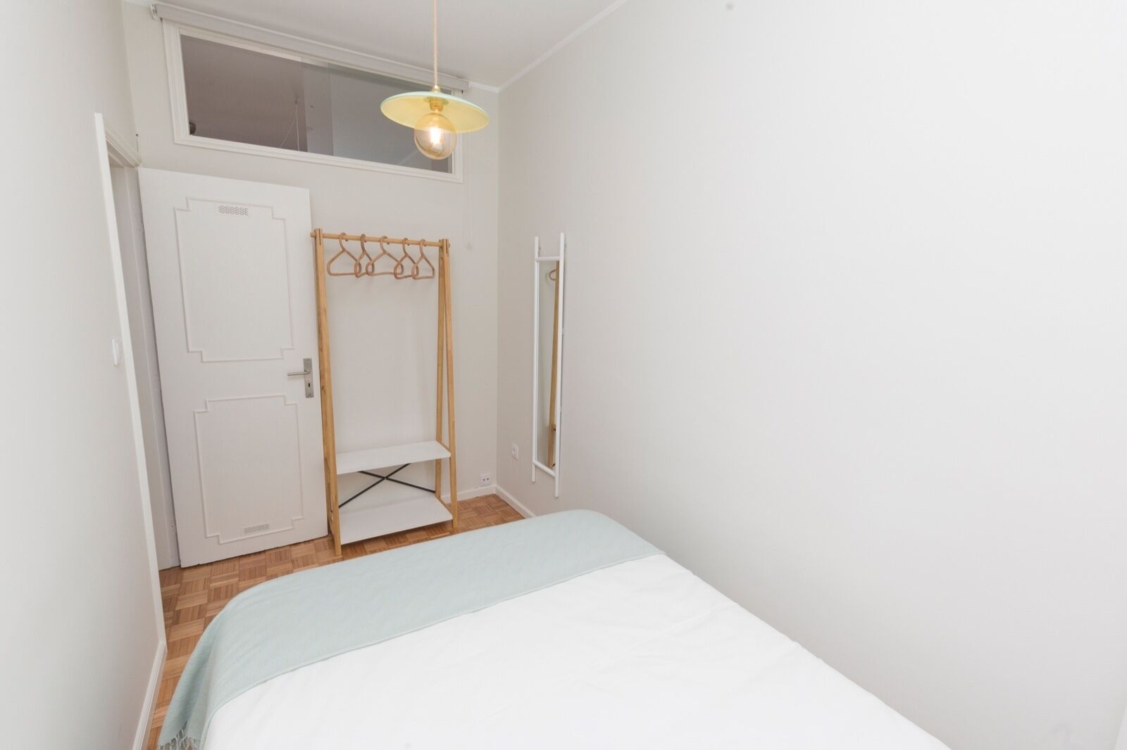 A small, bright room with a bed, a wooden hanger rack, a window, and a hanging light fixture.