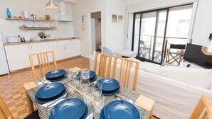 Une cuisine moderne avec des meubles blancs, une table à manger dressée avec de l’assiette et des verres bleus, et une grande fenêtre donnant sur un balcon.