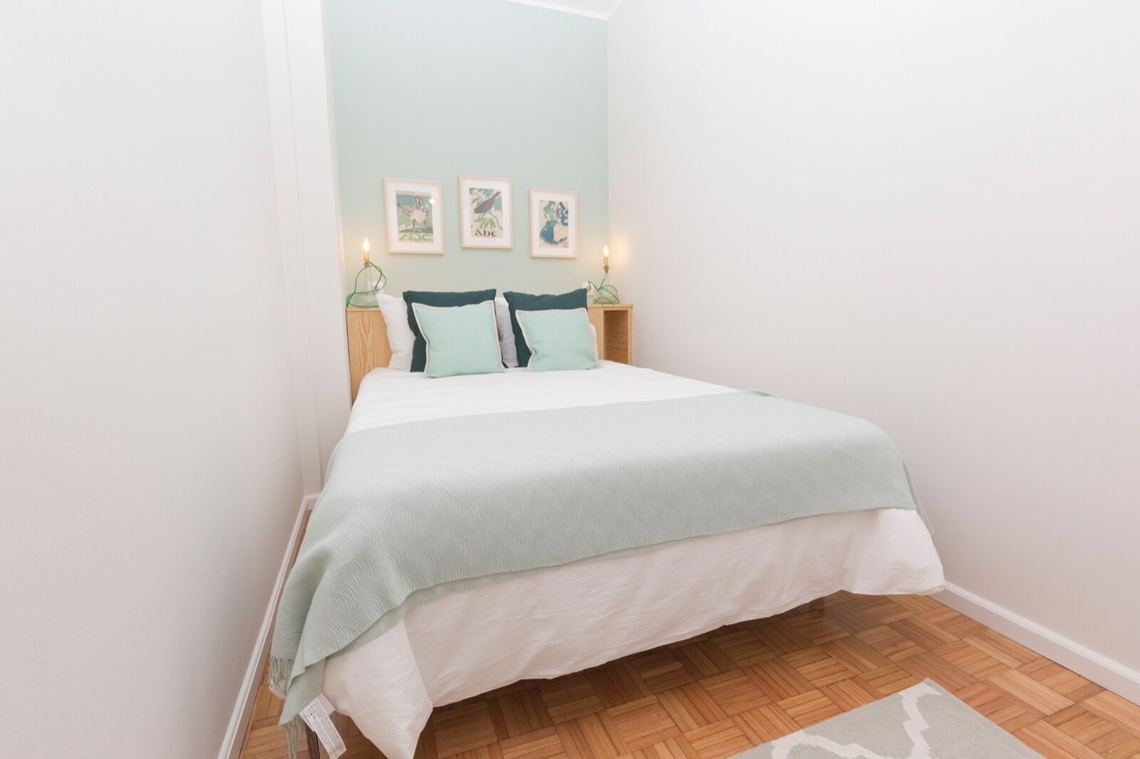 A neatly made bed with a light green bedspread and two pillows, a wooden nightstand with a lamp, and three framed pictures on the wall.