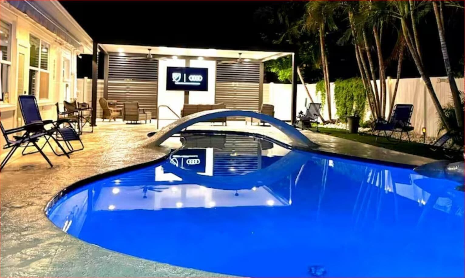 Una piscina con zona de descanso y un edificio con un letrero al fondo.