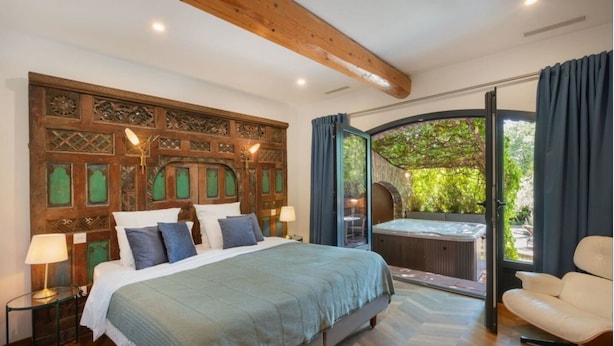 Ein Schlafzimmer mit einem großen Bett, einem Holzkopfende, einer Glastür, die zu einem Whirlpool führt, und Blick auf die grüne Landschaft.