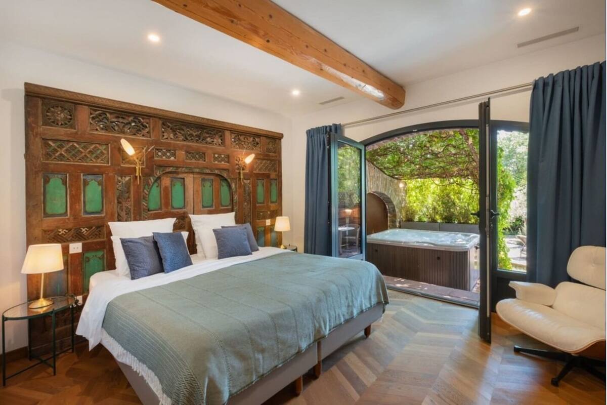 Een slaapkamer met een groot bed, een houten hoofdeinde, een glazen deur naar een jacuzzi en uitzicht op groen.