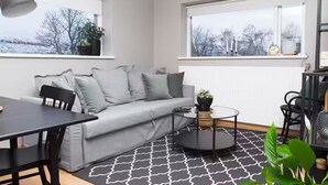 A modern living room with a grey sofa, black coffee table, and a dining table with chairs. There are two windows with views of trees and a cloudy sky. - Briet Apartments Akureyri, 2 bedroom (Akureyri)