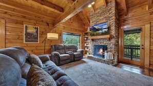 A cozy log cabin interior with a stone fireplace, leather sofas, and a large window overlooking greenery. - Bearly Behavin’ | Hot Tub, Pool, Arcade Game, MORE (Pigeon Forge)