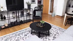 Ein Wohnzimmer mit einem schwarzen Fernseher auf einem Schrank, einem schwarzen Sessel mit Kissen, einem runden schwarzen Couchtisch mit Pflanze und einem gemusterten Teppich.