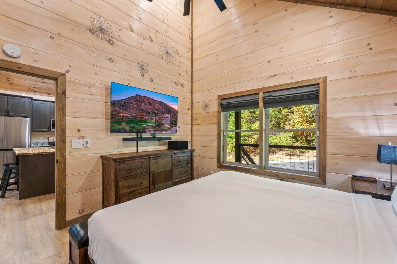 A wooden cabin bedroom with a bed, a dresser, a TV mounted on the wall, and a window with a view of greenery.
