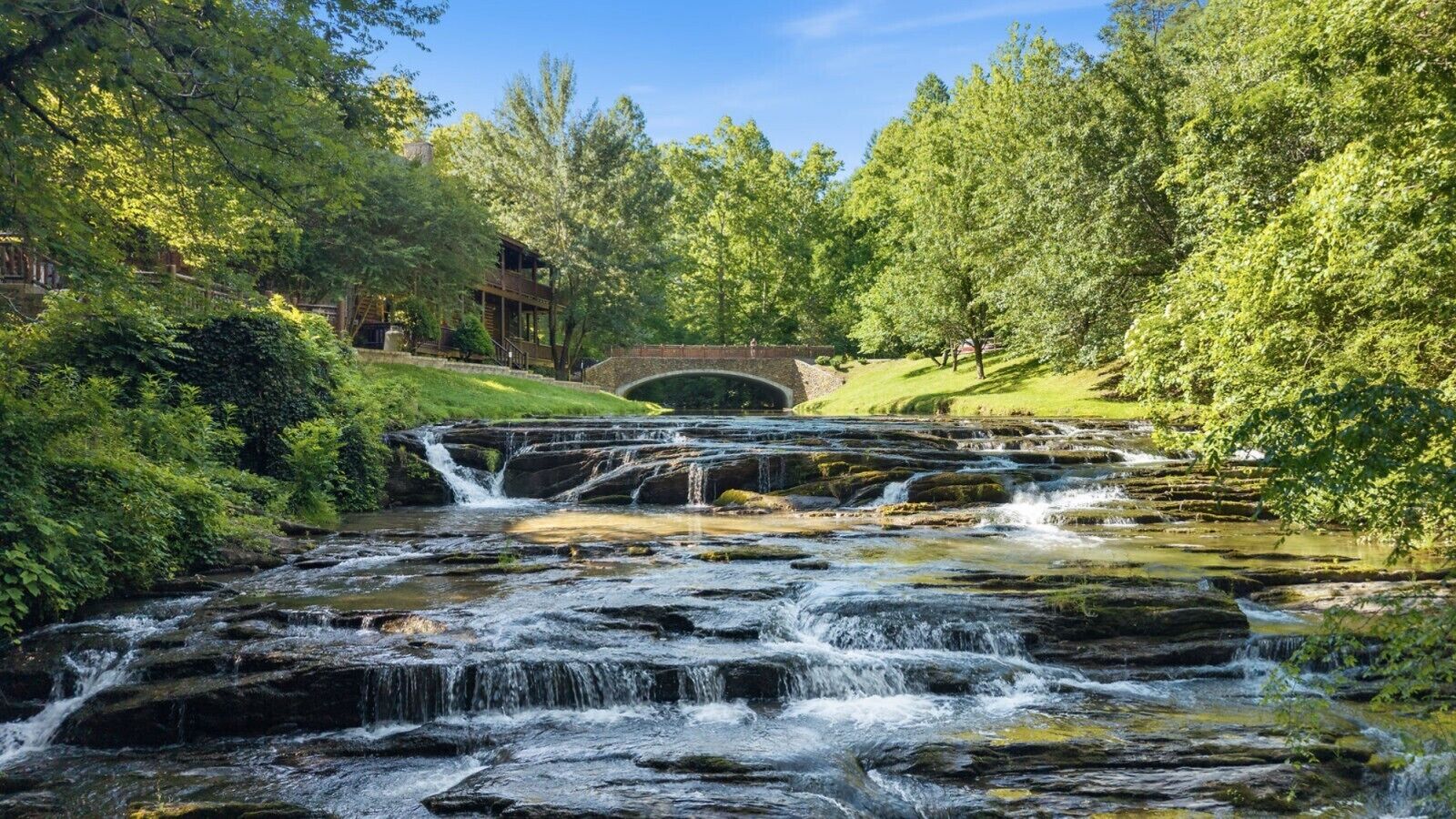 A natural landscape with a cascading waterfall, a wooden bridge, and lush greenery.