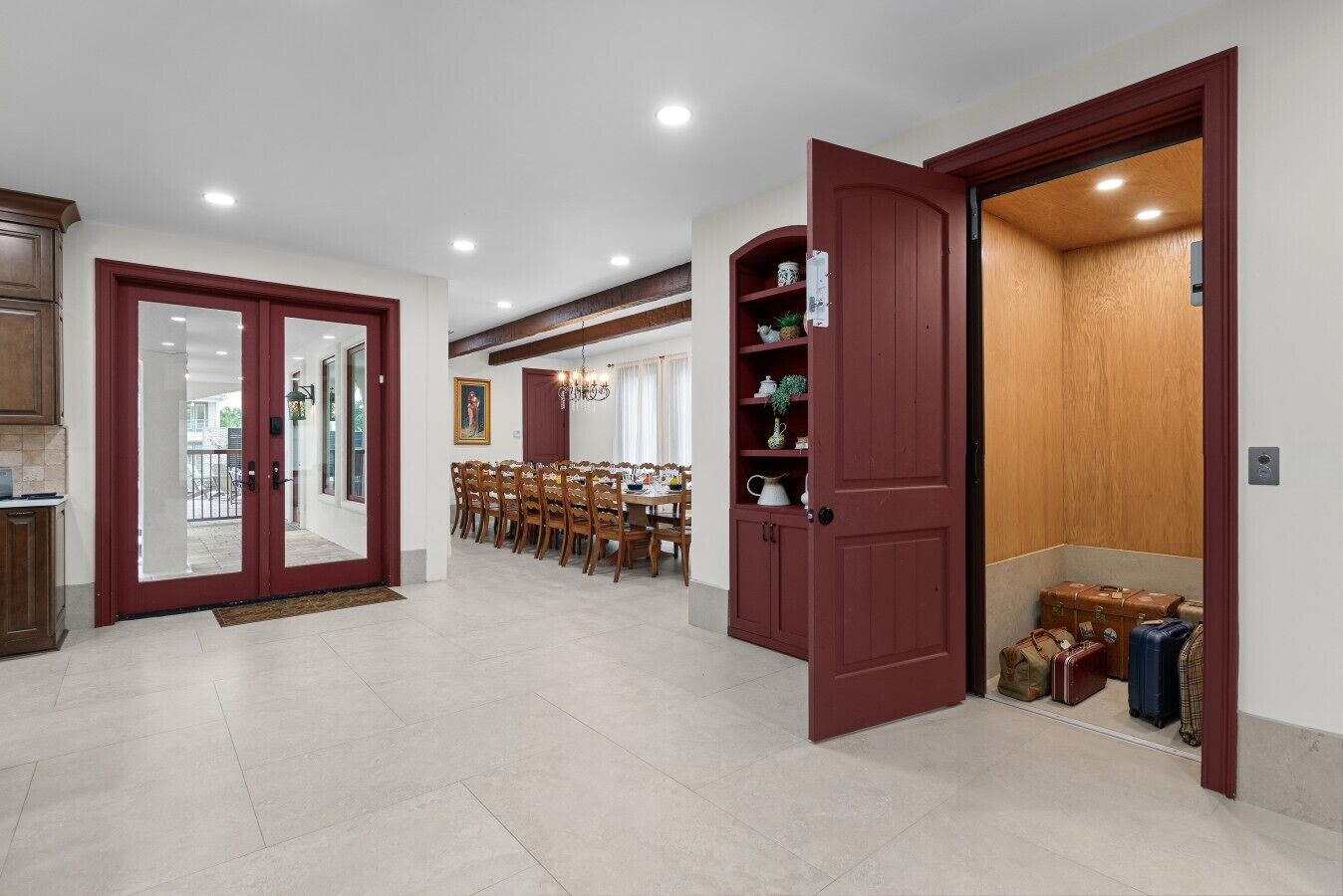 Un comedor amplio con una mesa larga y sillas, una puerta roja que da a un cuarto de almacenamiento con maletas, y vista al exterior por una puerta de vidrio.