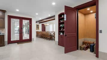 Un comedor amplio con una mesa larga y sillas, una puerta roja que da a un cuarto de almacenamiento con maletas, y vista al exterior por una puerta de vidrio.