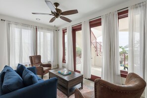 Ein Wohnzimmer mit Deckenventilator, einer Couch, einem Couchtisch mit einem Buch und Blick auf einen Balkon mit Geländer.