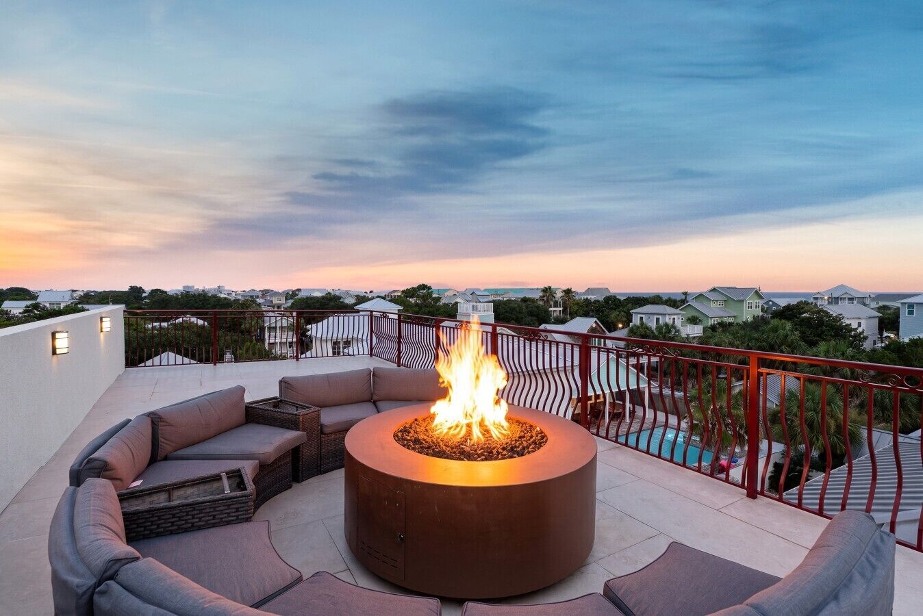 Una terraza en la azotea con un brasero circular rodeado de asientos acolchados, con vistas a una zona residencial y una piscina.