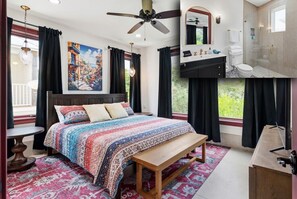 A bedroom with a bed, a bench, a ceiling fan, a window with curtains, a bathroom with a shower, and a painting on the wall.