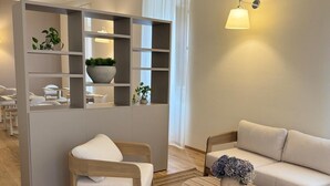 Un salon moderne avec un canapé, une table basse et un fauteuil. On y trouve une bibliothèque avec des plantes et une lampe.