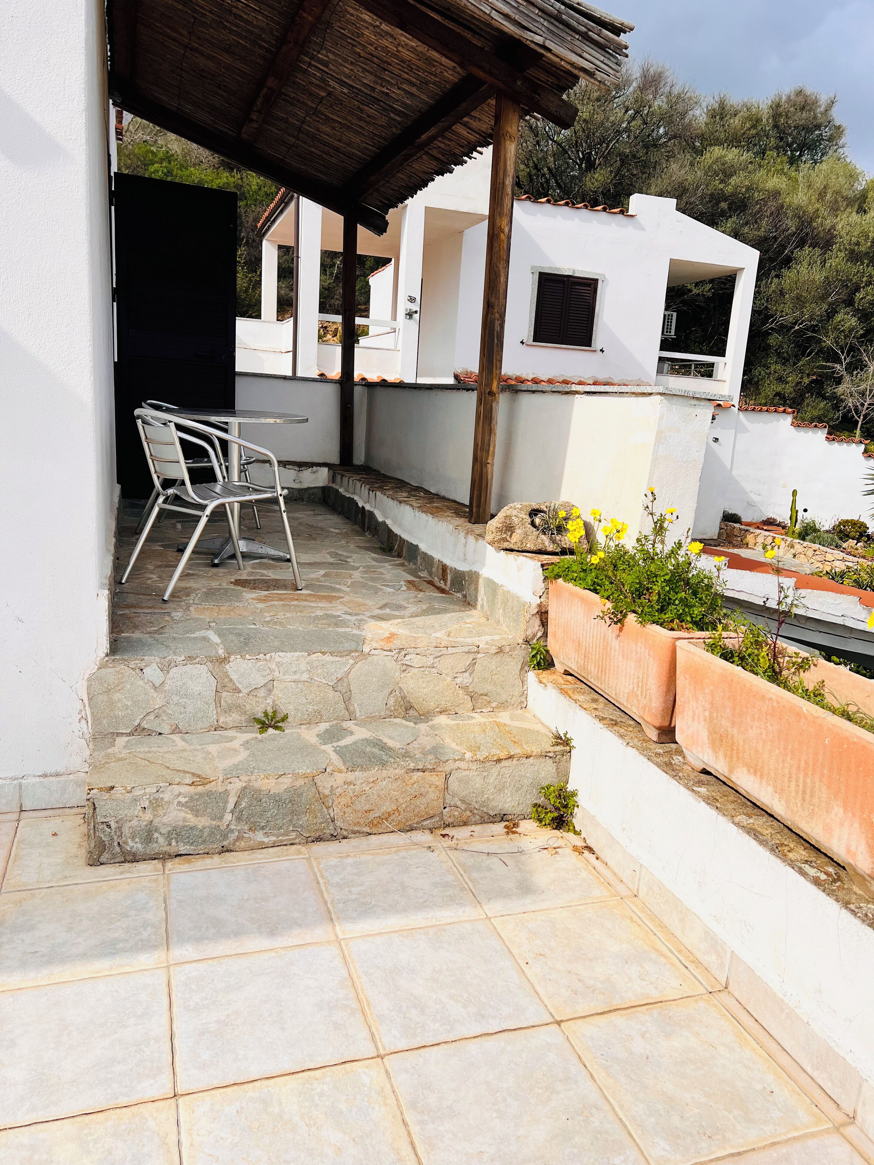 Un patio con escalones de piedra que conducen a un edificio blanco, una mesa y sillas de metal, y un pequeño jardín con plantas en macetas.