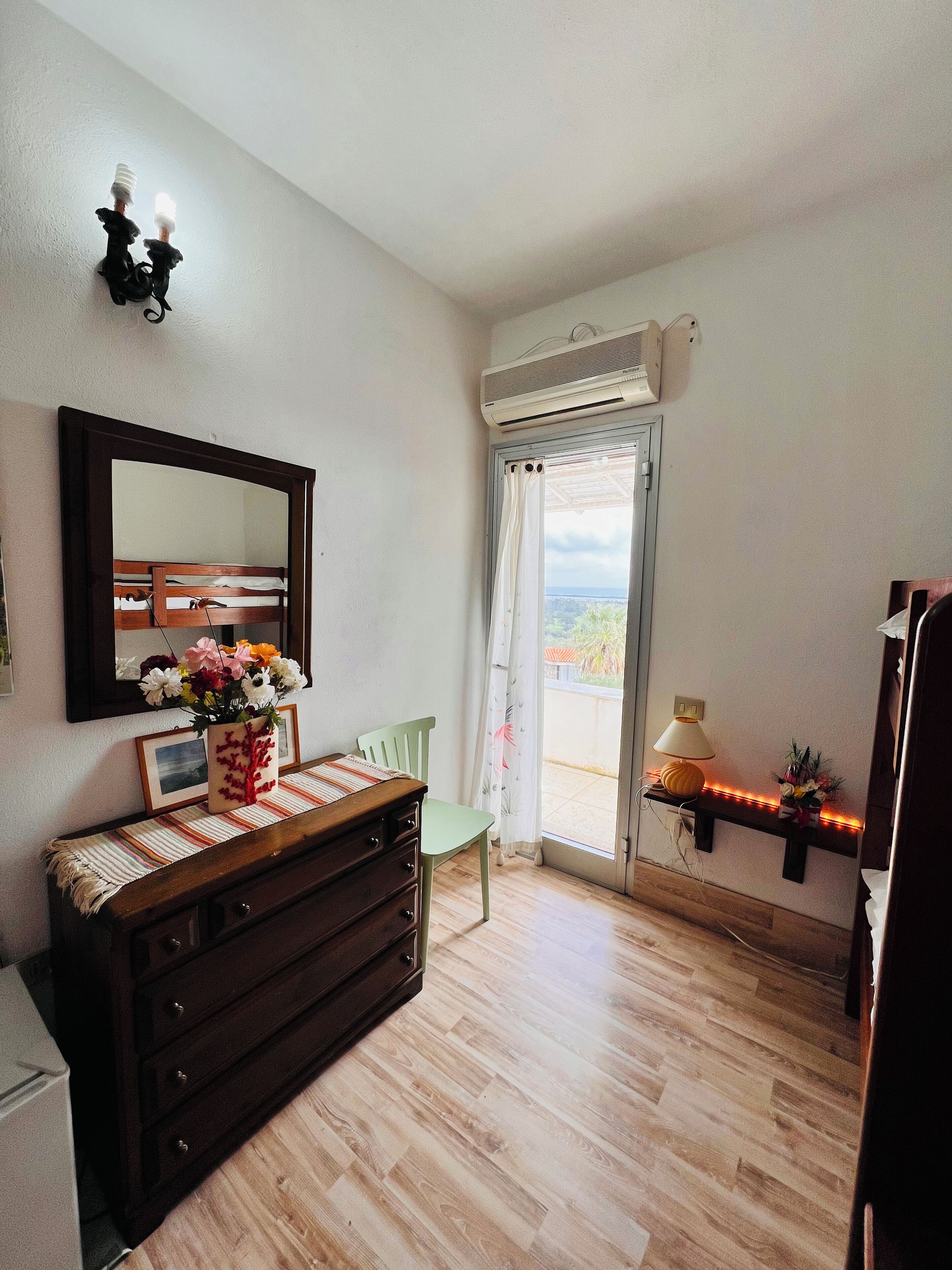 A room with a wooden dresser, a mirror, a window with curtains, a small table with a lamp, and a view of the outdoors.