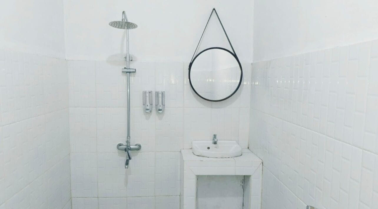 Une salle de bain avec des murs carrelés de carreaux blancs, un miroir rond, un lavabo et une douche équipée d’un pommeau de douche à main.