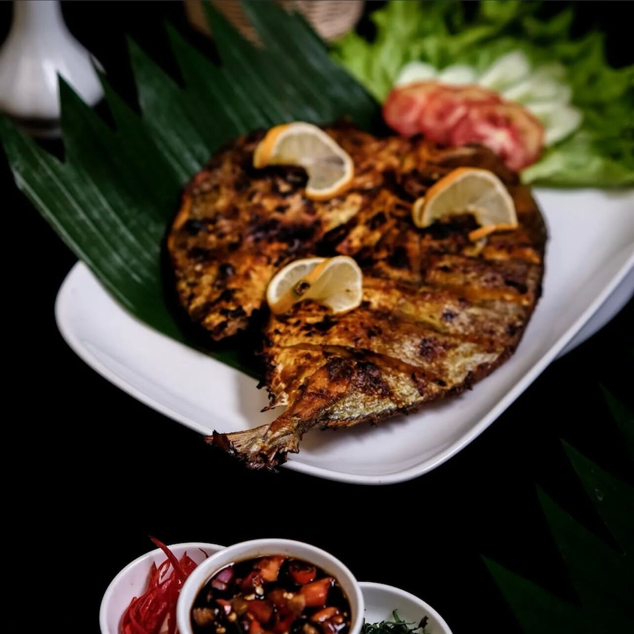 A grilled fish with lemon slices on a white plate, accompanied by a side of red chili sauce and a small bowl of dipping sauce.