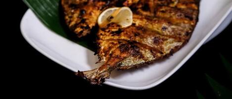 A grilled fish with lemon slices on a white plate, accompanied by a side of red chili sauce and a small bowl of dipping sauce.