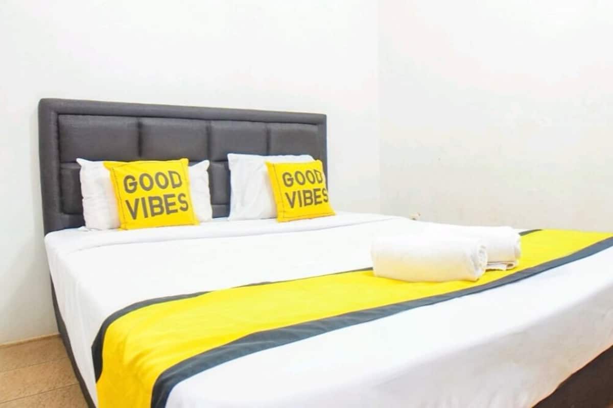 Egy szürke fejrésszel, fehér ágyneművel és két sárga párnával ellátott ágy, melyeken a „GOOD VIBES” felirat szerepel.