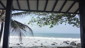 A beach view through a covered area with a palm tree and rocks. - La Plage Beach Club & Resort (Cikakak)