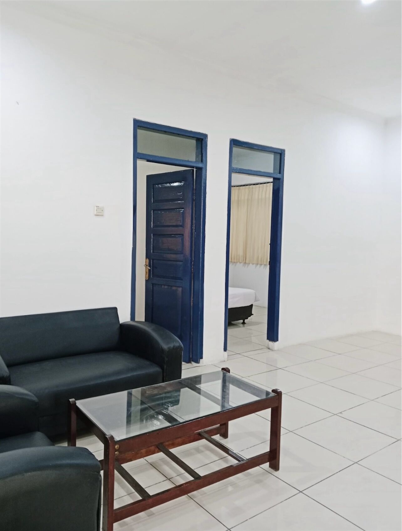 A room with a dark-colored sofa, a glass-top coffee table, and a bed with a white mattress and blue curtains.