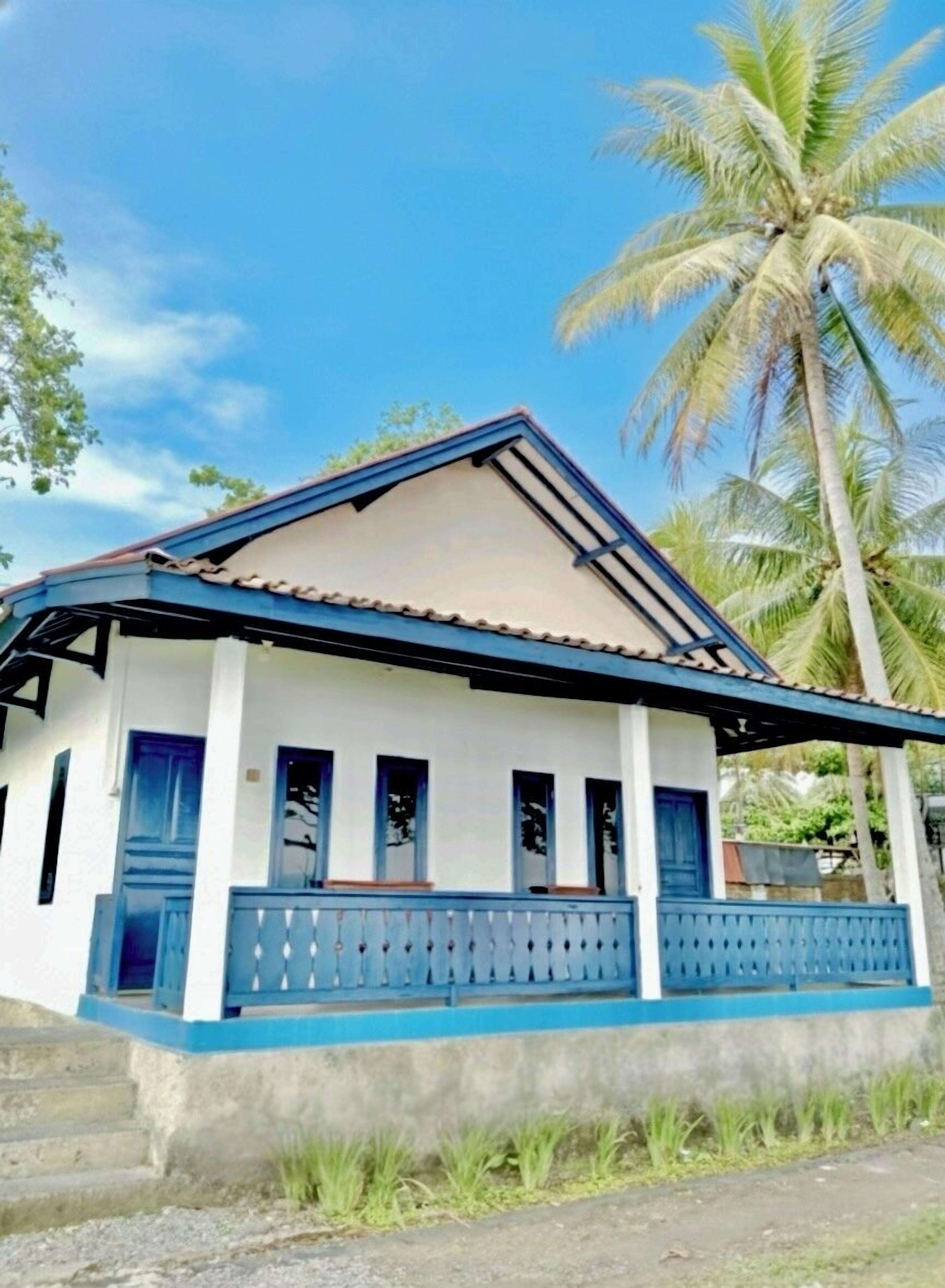 Una casa de dos pisos con fachada azul y blanca, tejado de tejas y un balcón con barandilla azul.