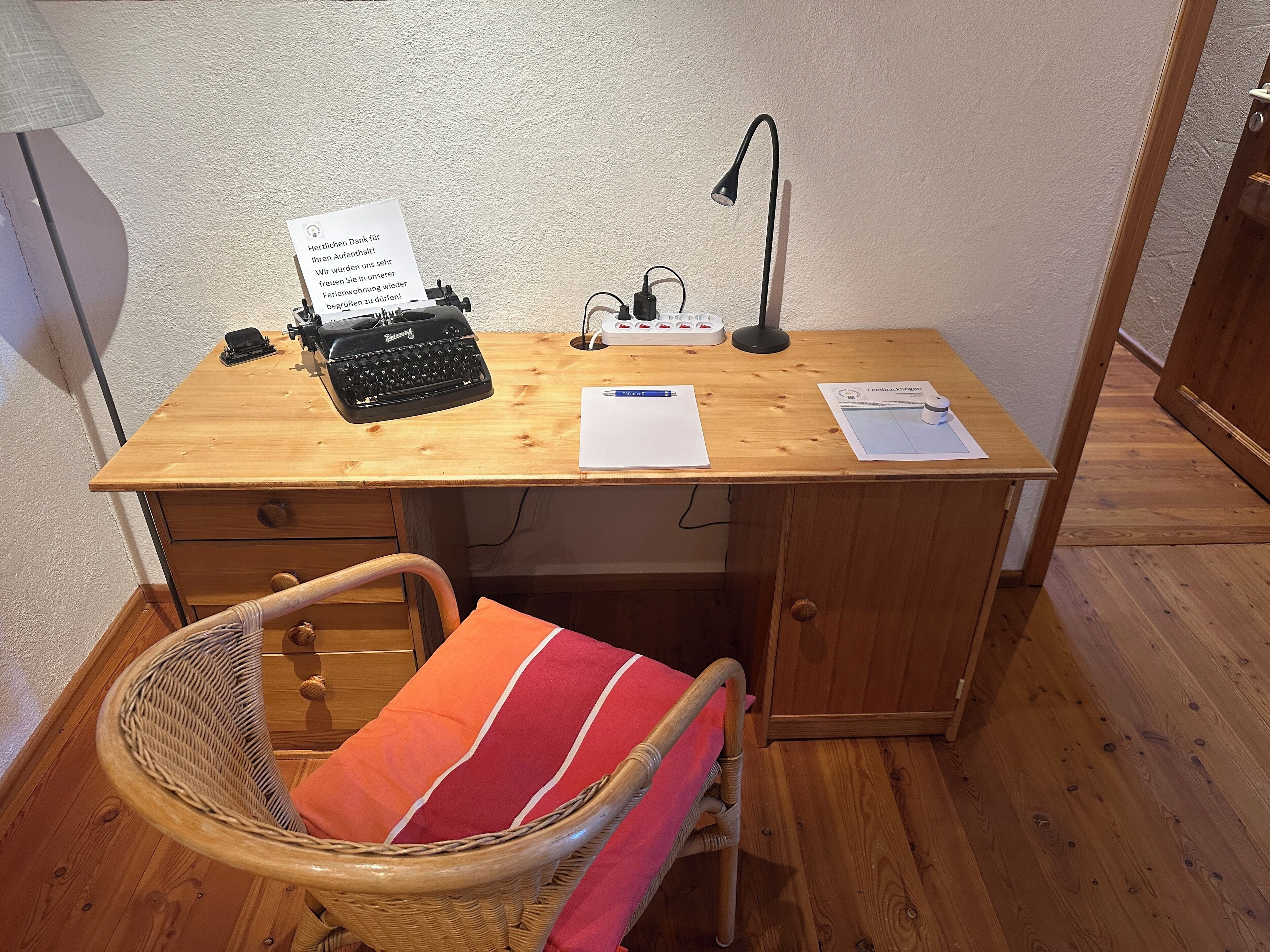 A wooden desk with a typewriter, lamp, papers, and a wicker chair with a cushion.