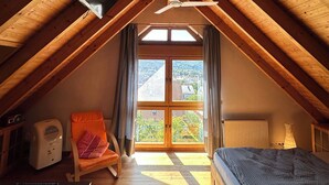 A cozy attic bedroom with a sloped wooden ceiling, a ceiling fan, a large window with curtains, a bed, a chair, a heater, and a TV mounted on the wall. - FEWOBuergstadt.de Light-flooded, cozy, spaciousFeWo with distant view (Bürgstadt)