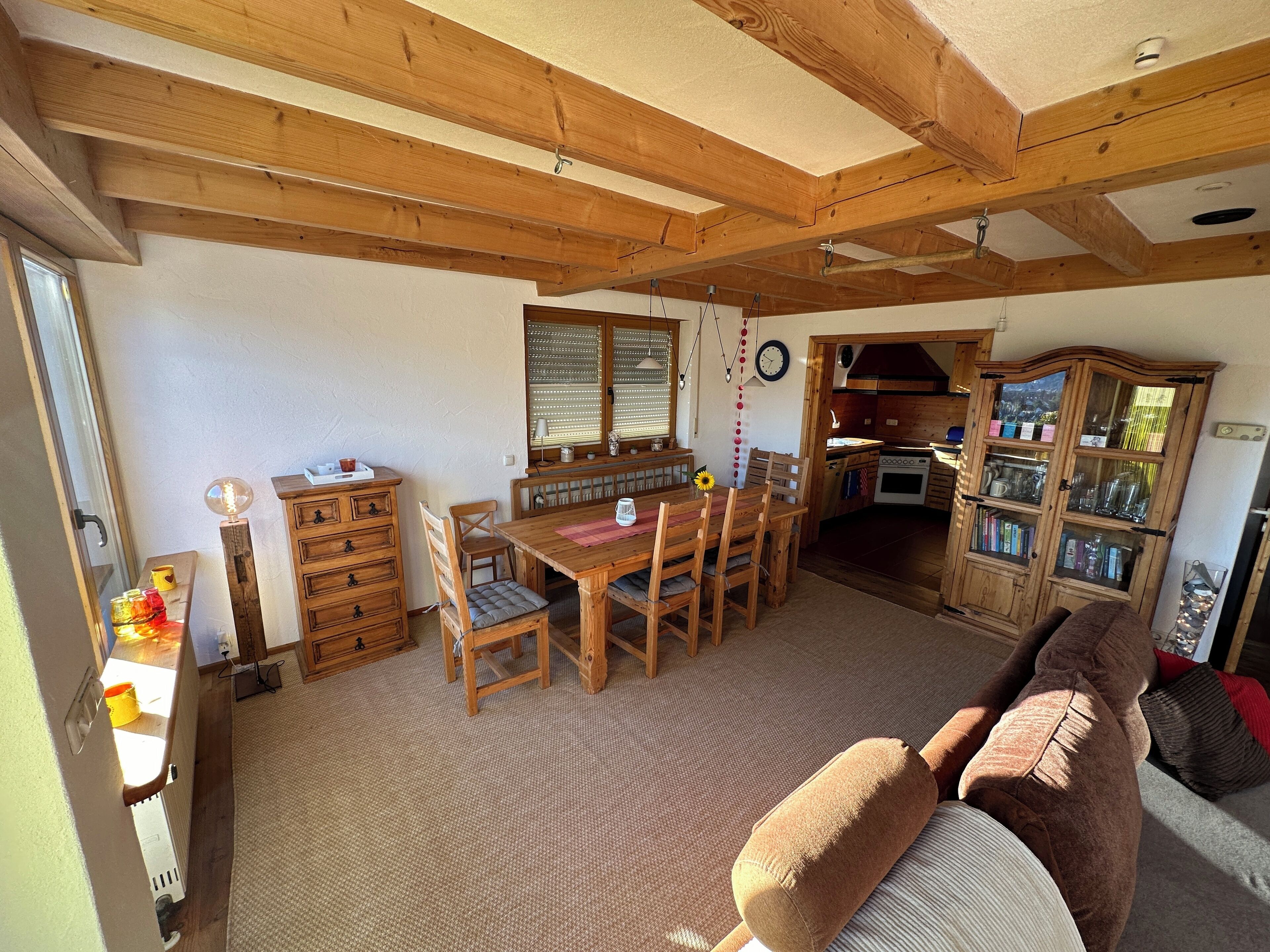 A cozy living room with a wooden dining table, chairs, a sofa, and a kitchen area with cabinets and a stove.