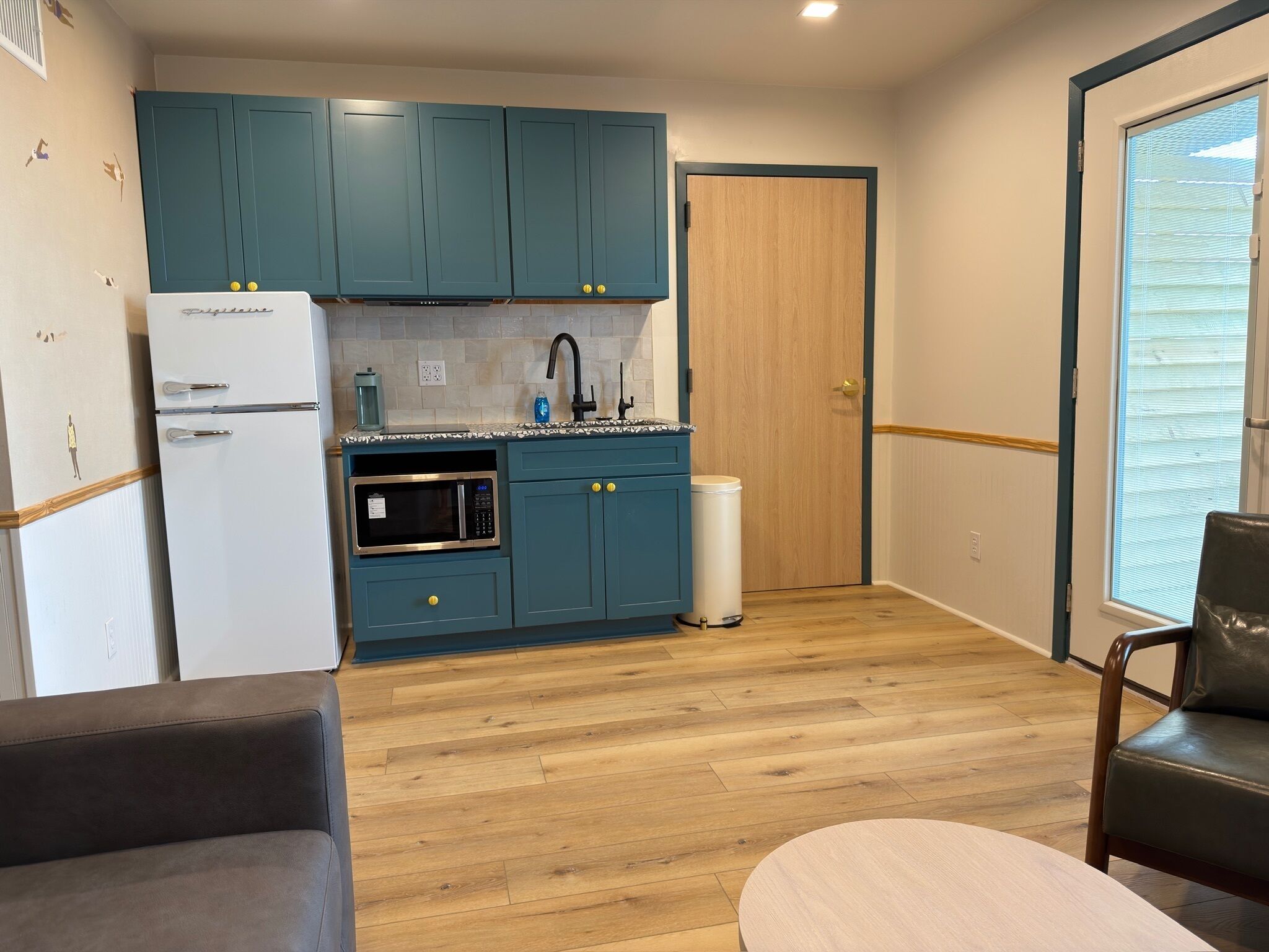 A compact kitchen with teal cabinets, a white refrigerator, a microwave, and a sink. There's a trash bin and a wooden door.