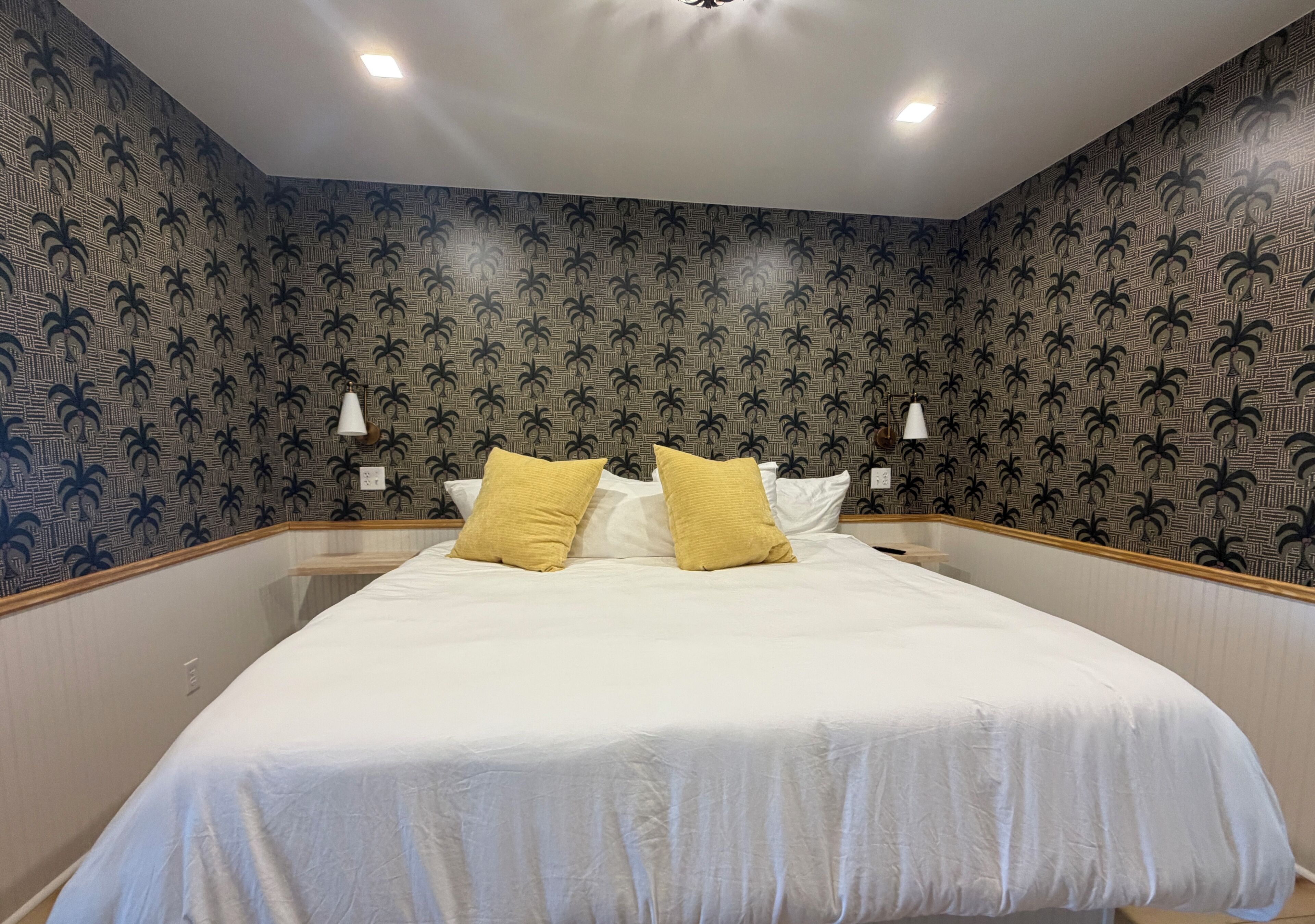 A neatly made bed with white and yellow pillows, a wooden headboard, and a wall with a palm tree pattern.
