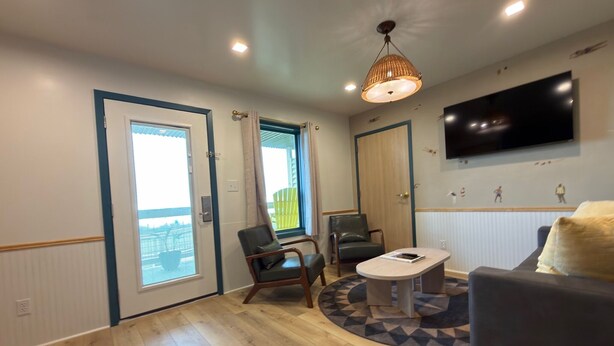 A room with a wooden floor, a round table, two chairs, a sofa, a TV mounted on the wall, and a door with a view to the outside. - The Breeze Beachfront Resort (Surfside Beach)