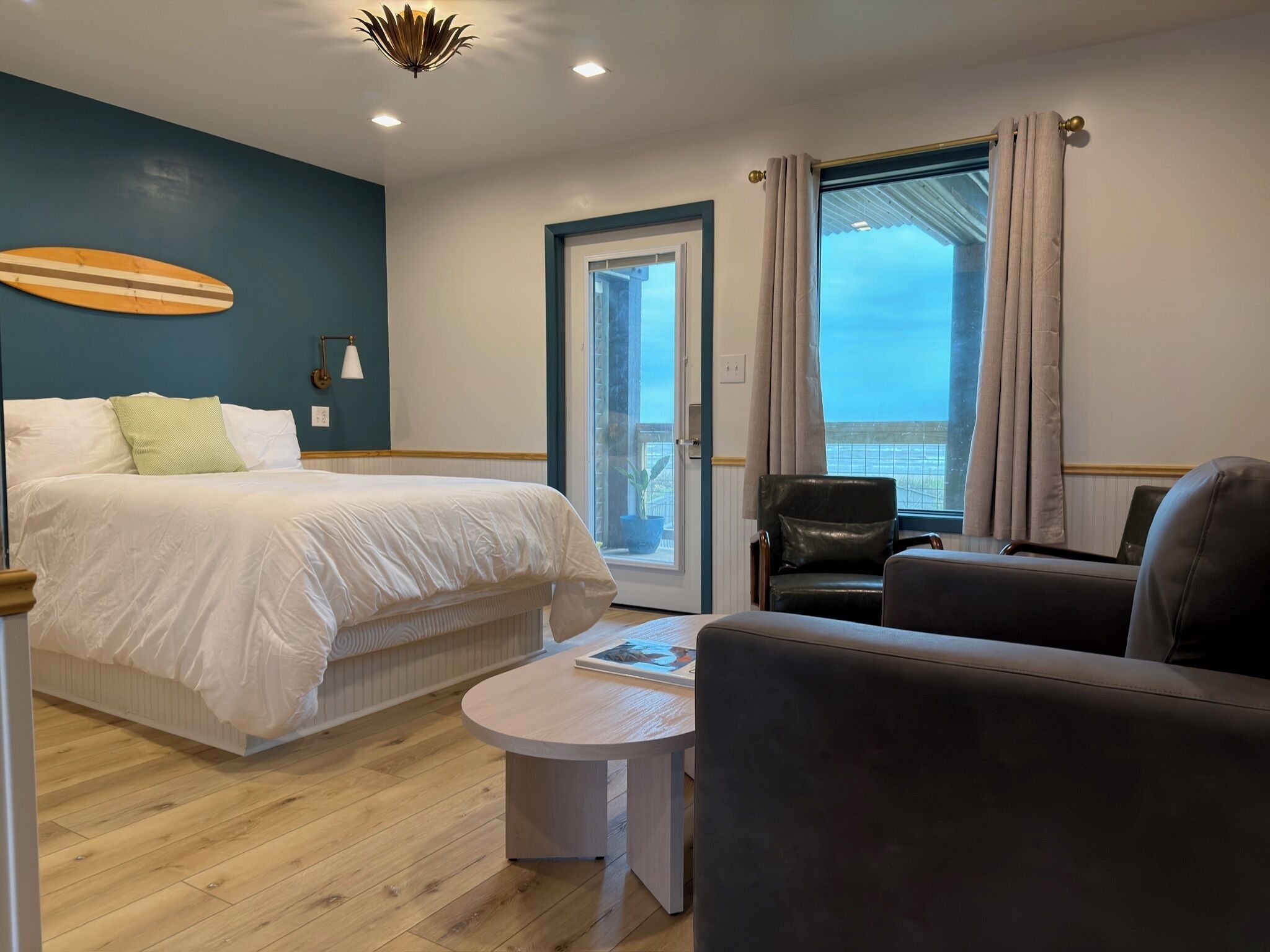 A hotel room with a bed, a round table, a sofa, a chair, a potted plant, and a view of the sea through a window.